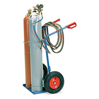 a broad blue FETRA® steel bottle trolley made of steel tubes, with on three sides skirted loading shovel, air tyres on red rims, red handles on back-bent push bars and chain-fastening for the gas bottles, and loaded with two upright gas bottles with pressure minimizers and hosing, isolated on white background