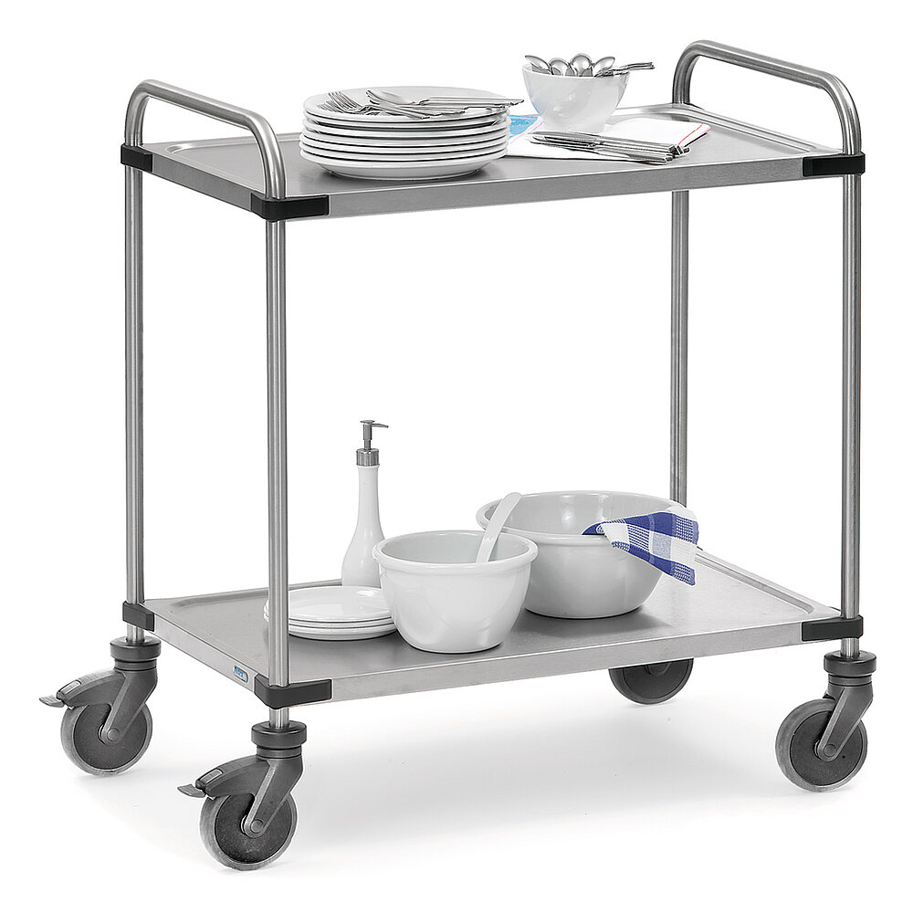 a silvery-shining FETRA® trolley made of stainless steel tube, with two storeys, each with lightly depressed stainless steel sheets and four steering wheels, two of which are fastenable, and loaded with stacks of plates, cutlery and hospitality-related articles, isolated on white background