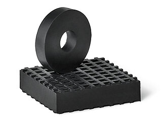 a black, square rubber board made of elastomer NBR with small square indentions on the surface as non-slip protection, on top of which stands an upright black, round rubber disc without surface profiling and a centered hole, isolated on white background 