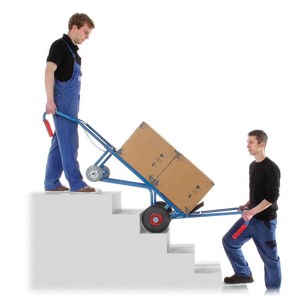 two workers on a grey concrete staircase in front of and behind a blue FETRA® appliance truck made of steel tube with black tyres on red rims, additional small supporting wheels at the rear side, a large brown cardboard box placed on the broad loading fork and additional carry-bars with red handles, isolated on white background