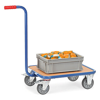 a blue FETRA® dolly with gooseneck handle made of steel tube, with light-brown, low-level wooden loading platform, two fixed wheels at the front, two fastenable steering wheels at the rear, a long orthogonal push bar with red handle, and loaded with a grey plastic box with orange lashing straps, isolated on white background