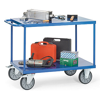 a blue FETRA® table top cart made of steel tubes and profiled steel, with two storeys, push bar, flush-fitting sheet metal platforms, two fixed wheels at the front, two fastenable steering wheels at the rear, and loaded with power tools and a hydraulic cylinder, isolated on white background