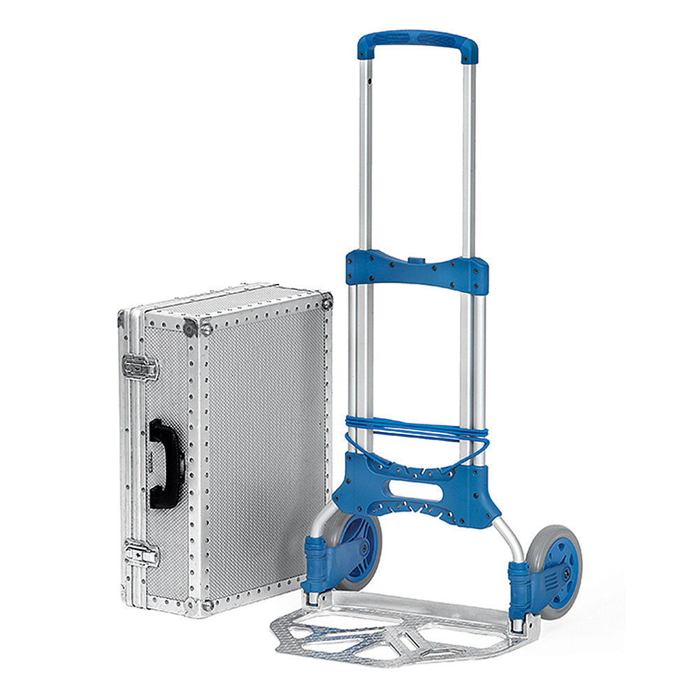 a collapsible FETRA® compact hand truck made of silvery aluminium tubes with blue plastic handle, unfoldable broad loading shovel and elastic rubber fastening strap, next to it an upright aluminium suitcase, isolated on white background